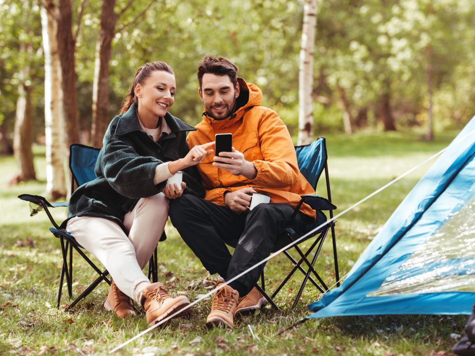 camping, tourism and travel concept - happy couple with smartphone drinking tea at campsite